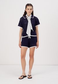 Completo corto in maglia a rete blu navy con rifiniture bianche, composto da una maglia con colletto e maniche corte e pantaloni corti, accentuato da una sciarpa a pois annodata al collo.