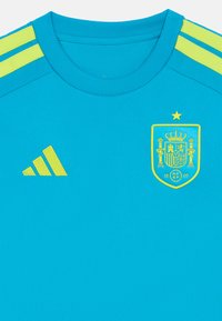 Camisola atlética turquesa com detalhes amarelos, apresentando um decote redondo, três riscas amarelas em cada ombro e o brasão da Espanha com uma estrela dourada.