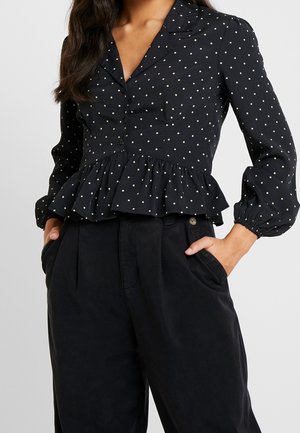 Frau trägt eine schwarze Bluse mit weißen Punkten und Rüschensaum sowie eine hoch taillierte schwarze Hose, die Hände in den Taschen.