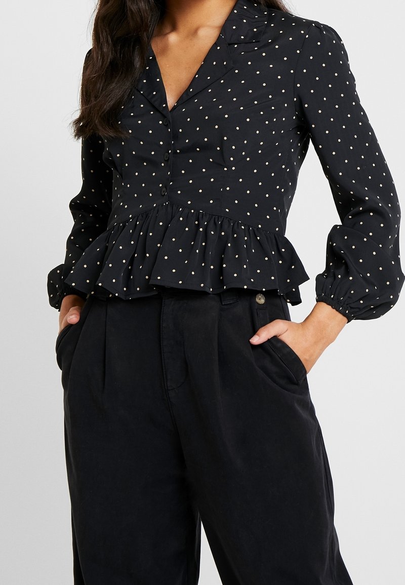 Femme portant un chemisier noir à boutons avec des pois blancs et un ourlet à volants, assorti à un pantalon noir taille haute, les mains dans les poches.