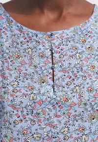 Blouse bleu clair avec motif floral en rose, vert et blanc. Présente un col rond et trois boutons décoratifs à l'avant.