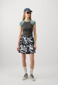 Top de manga corta gris y verde azulado, pantalones cortos con patrón en blanco y negro, zapatillas grises, calcetines blancos con texto y una gorra de mezclilla.
