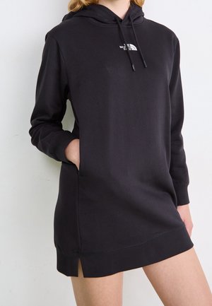 Persona que lleva un vestido con capucha de manga larga negro con un bolsillo frontal y el logo de The North Face en el pecho, de pie frente a un fondo blanco.