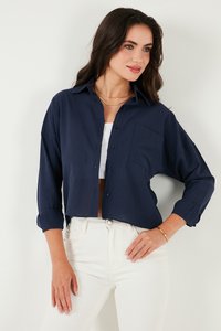 Navy blue button-up shirt with a relaxed fit, dropped shoulders, and a front pocket, made of soft fabric. Worn over a white top and white jeans.
