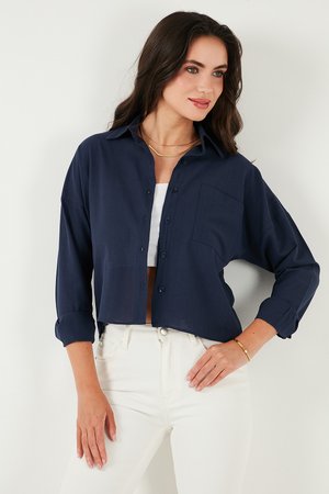 Navy blue button-up shirt with a relaxed fit, dropped shoulders, and a front pocket, made of soft fabric. Worn over a white top and white jeans.