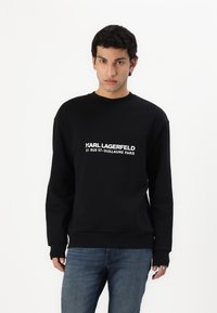 Crna dukserica s okruglim izrezom, na kojoj se nalazi bijeli logotip s natpisom: "KARL LAGERFELD 21 RUE ST-GUILLAUME PARIS." Izrađena od mekanog pamučnog materijala.