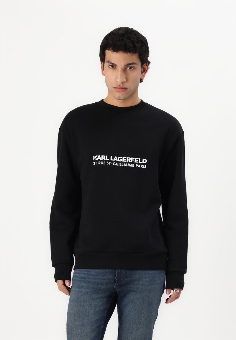 Crna dukserica s okruglim izrezom, na kojoj se nalazi bijeli logotip s natpisom: "KARL LAGERFELD 21 RUE ST-GUILLAUME PARIS." Izrađena od mekanog pamučnog materijala.