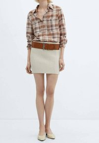 Persona che indossa una mini gonna a coste beige, una cintura marrone, scarpe basse beige e una camicia a quadri marrone con maniche lunghe arrotolate.
