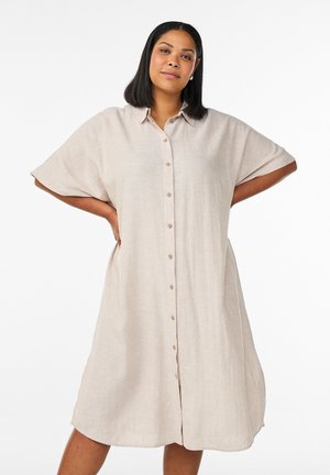 Woman with medium skin tone and shoulder-length dark hair wears a beige button-up loose dress, standing with hands on hips against white background.