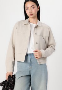 Veste en velours côtelé beige avec un col, fermeture par boutons à l'avant et texture ribbed. Associée à un jean taille haute bleu clair.
