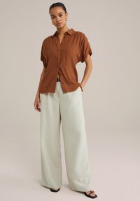 Camicia marrone a maniche corte con un motivo testurizzato, abbinata a pantaloni larghi verde chiaro e sandali neri. Gli accessori includono un bracciale d'oro.