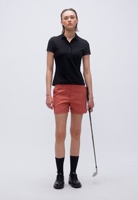 Γυναίκα που στέκεται κρατώντας ένα μπαστούνι γκολφ, φορώντας μαύρο πόλο με κοντά μανίκια, κόκκινα σορτς, μαύρες κάλτσες και μαύρα παπούτσια, μπροστά σε απλό φόντο.