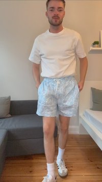 Lichtblauwe zwemshorts met een wolkenpatroon, elastische tailleband met trekkoord, gecombineerd met een wit T-shirt en witte sokken, op een houten vloer.