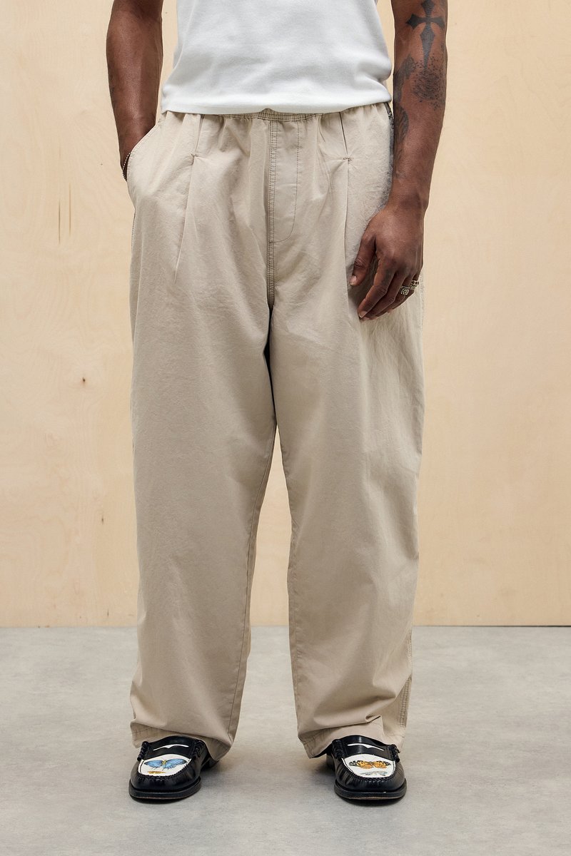Beige, plisserede bukser lavet af letvægtigt stof; løs pasform. Båret med sorte sko med dekorative detaljer. Enkel, funktionel design.