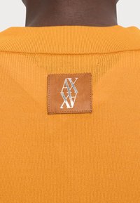 Oranssi neulepaita, jossa on teksturoitua kangasta. Sisältää ruskean nahkapatchin, jossa on kohokuvioitu logo takaneulassa.