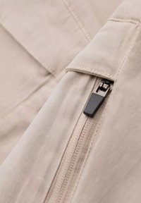 Gros plan sur un tissu beige montrant une poche zippée avec une tirette noire. Le textile a une texture lisse et des détails de couture subtils.