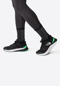Scarpe da corsa nere con accenti verdi, caratterizzate da una tomaia in rete e un'intersuola bianca, abbinate a calze nere a strisce e leggings neri.
