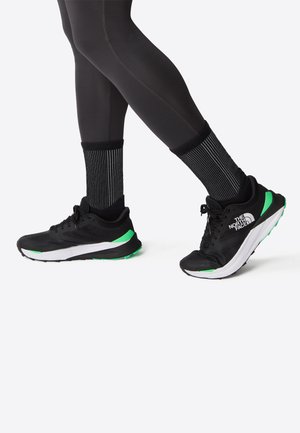 Baskets de course noires avec des accents verts, dotées d'une tige en mesh et d'une semelle intermédiaire blanche, associées à des chaussettes noires rayées et des leggings noirs.