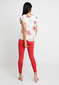 Blusa bianca con motivo floreale e maniche arricciate, abbinata a jeans rossi aderenti. Il modello è in piedi con le spalle rivolte verso la macchina fotografica, indossando tacchi nudi.