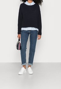 Pull en maille bleu marine porté par-dessus une chemise rayée bleu clair, associé à un jean bleu et des baskets blanches, tenant un petit sac à main noir.