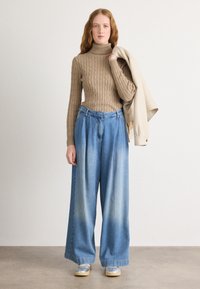 Un pull col roulé en tricot beige, un jean taille haute à jambes larges bleu clair et un vêtement beige drapé sur un bras. Des baskets gris clair complètent le style.