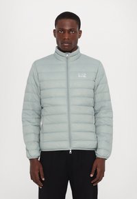 Light blue puffer jacket with quilted stitching, high collar, and zip closure. Features a small logo on the left chest and elastic cuffs.