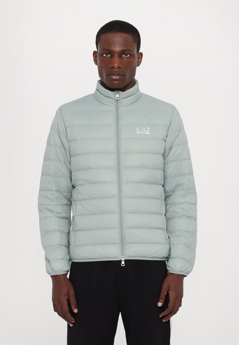Light blue puffer jacket with quilted stitching, high collar, and zip closure. Features a small logo on the left chest and elastic cuffs.