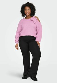 Rosa gerippter Pullover mit einer Schleifendetail auf der linken Schulter, kombiniert mit schwarzen Schlaghosen und schwarzen Schuhen. Lockiges Haar, lächelnd.