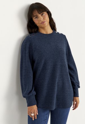 Kvinde med mørkt hår iført en navyblå langærmet sweater med knapdetaljer på skuldrene og blå jeans, der står op ad en hvid væg.