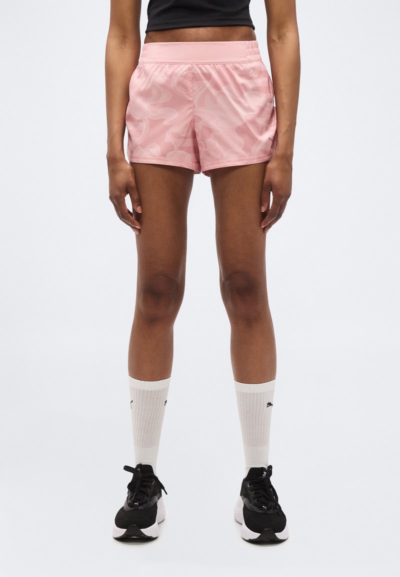 Νέος που φοράει ροζ σορτς με σχέδια, λευκές κάλτσες μέχρι το γόνατο και μαύρα αθλητικά παπούτσια, στέκεται μπροστά σε λευκό φόντο.