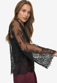 Zwarte kantblouse met lange, uitlopende mouwen, voorzien van een ingewikkeld bloemenpatroon en een transparante textuur. Gepaarde met bordeauxrode broeken.