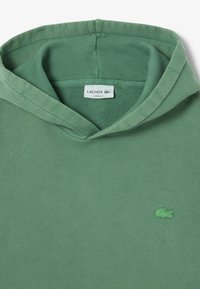 Lockere grüne Kapuzenjacke aus Baumwolle. Verfügt über eine verstellbare Kapuze, einen gerippten Halsausschnitt und ein kleines gesticktes Logo auf der Vorderseite.