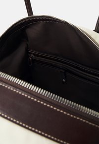 Beżowe wnętrze torby z ciemnobrązową tkaniną. Posiada kieszeń na zamek błyskawiczny i szczegóły przeszycia. Zamek błyskawiczny na górnej krawędzi.
