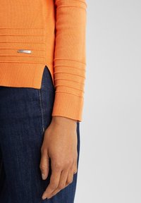 Oranges Sweatshirt mit texturierten Rippdetails an den Ärmeln, mit einem seitlichen Schlitz. Kombiniert mit dunkelblauen Jeans. Silbernes Markenlabel sichtbar.