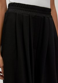 Zwarte geplooide broek met een elastische tailleband. De stof heeft een gladde textuur en een gestructureerd ontwerp, geschikt voor zowel casual als formele kleding.