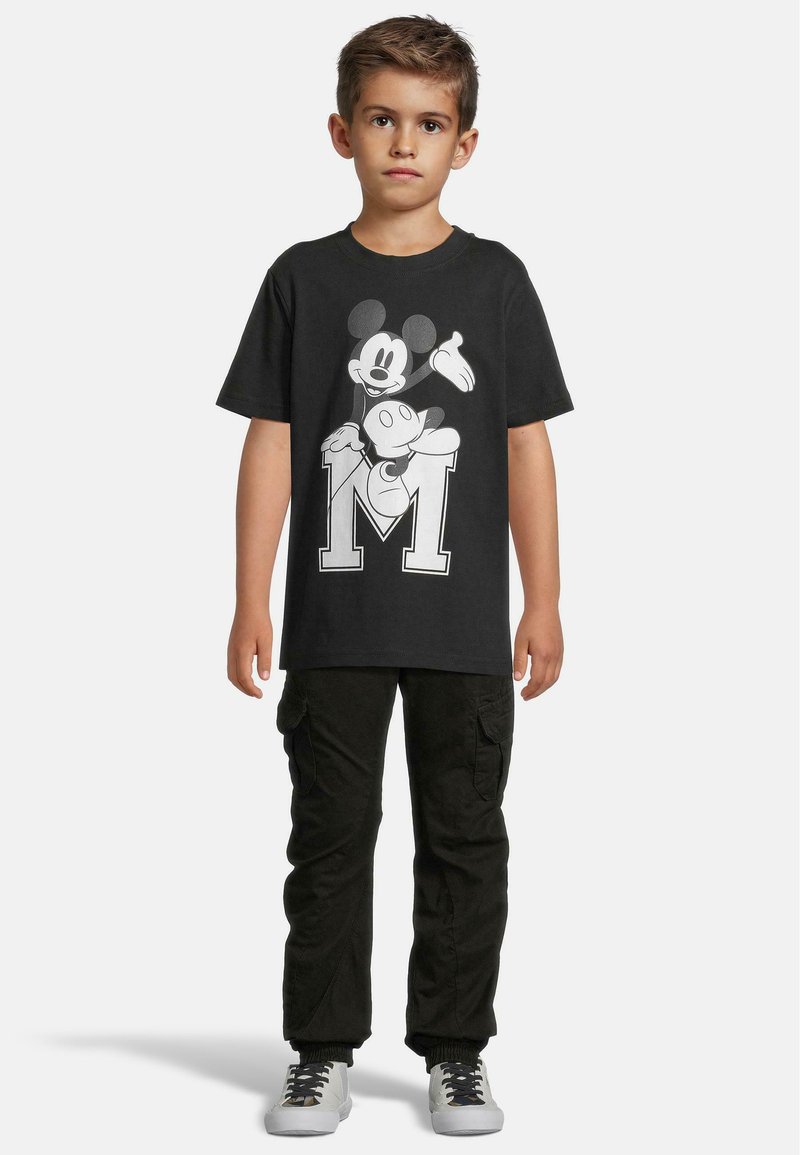 Černé bavlněné tričko s potiskem Mickey Mouse a bílým „M“ pod ním. Dítě oblečené v černých cargo kalhotách a šedých teniskách.