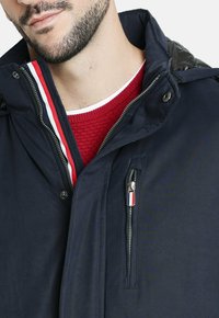 Chaqueta de marina con cremallera en el frente, un cuello interior de punto rojo y bolsillos laterales con un pequeño acento tricolor en el tirador de la cremallera.