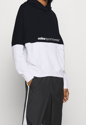 Osoba ubrana w czarno-białą bluzę Nike Sportswear z kapturem oraz czarne spodnie z białym paskiem z boku, ręka w kieszeni, na jednolitym tle.