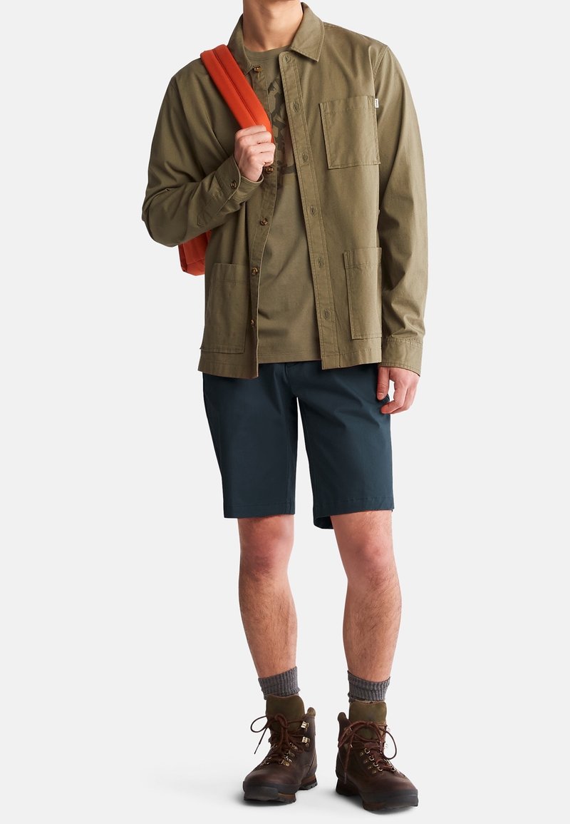 Personne portant une veste-chemise vert olive sur un t-shirt, un short marine, des chaussures de randonnée marron avec des chaussettes grises, et tenant une sangle de sac à dos orange.