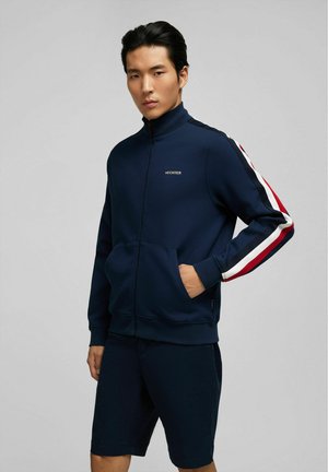 Navy zip-up jacket with white and red striped accents on sleeves, soft fabric, front pockets, and a small logo on the chest.