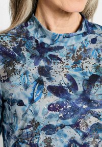 Bluse mit floralem Muster, gekennzeichnet durch einen Mock-Neck, überwiegend blau mit dunklen Akzenten, und einem strukturierten Finish mit silbernen Highlights.