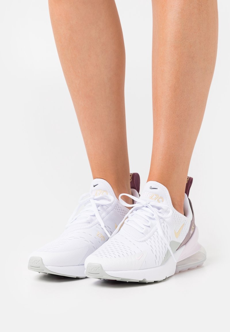 Jambes portant des baskets blanches Nike Air Max 270 avec des semelles gris clair et des accents violets sur fond blanc.