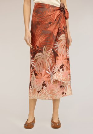 Frau trägt einen tropischen Wickelrock in Rost und Pink mit Blumenmuster, der an der Taille gebunden ist, dazu braune verzihrte Ballerinas.