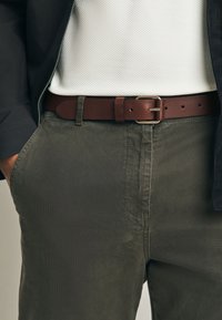 Cintura in pelle marrone con fibbia d'argento, indossata sopra pantaloni verde oliva con finitura strutturata e una maglietta bianca con texture.