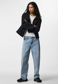 Jeune femme aux cheveux bouclés portant une veste noire, une chemise blanche, un jean bleu clair et des baskets noires, debout, les bras croisés.