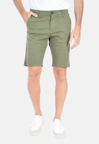 Pantaloni corti in cotone verde oliva con chiusura frontale a bottone, tasche laterali e design a gamba dritta, indossati con sneakers bianche.