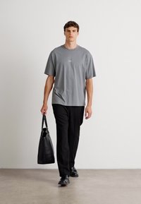 Grå bomull t-skjorte med en liten logo, kombinert med sorte bukser og en svart tote bag. Modellen har på seg svarte sko og står innendørs.