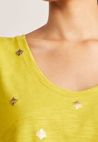 Žlutá krátkosrstá košile se širokým výstřihem, s ozdobnými detaily zlatého a hnědého vyšívání ve tvaru mušlí na přední straně. Hladká textura.