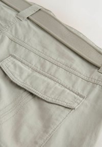 Zbliżenie na kieszeń spodni cargo z jasnoszarego materiału z klapką i przeszyciami oraz z pętlą na pasek z tego samego materiału nad nią.