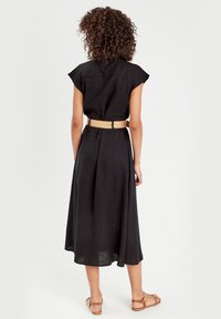 Femme aux cheveux bouclés portant une robe midi noire à manches courtes, une ceinture beige et des sandales plates marron, debout de dos.